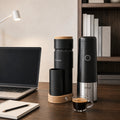 A coffee-making setup on a wooden desk featuring a black and wood coffee grinder, a black and stainless steel coffee maker, and a small cup of espresso, with a laptop, notebook, and bookshelf in the background.