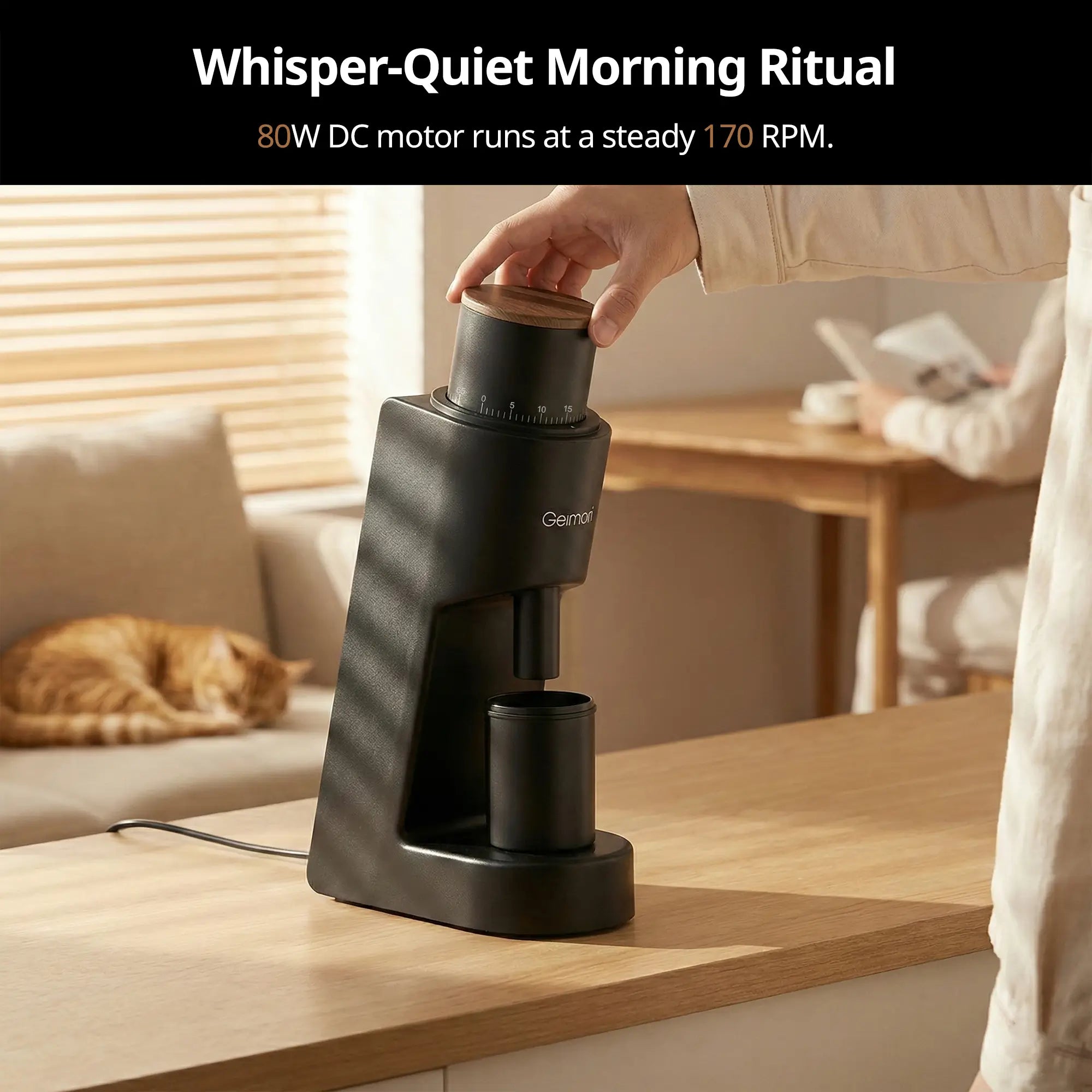 A black Geimori GU38 coffee grinder sits on a wooden countertop in a bright, modern kitchen. A hand is shown turning the top dial of the grinder.